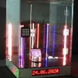 Beleuchtetes elektronisches Gerät im Glaskubus auf einem Riffelblechsockel. Ein rotes LED-Display zeigt das Datum "24.06.2024". Im Inneren sind rosa und blaue Leuchtröhren sowie eine lila Platine sichtbar.